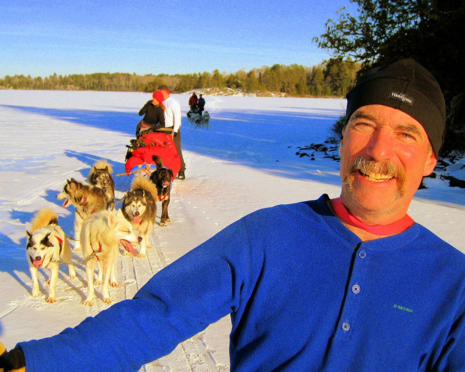 Man smiling with sled dogs pulling family behind him