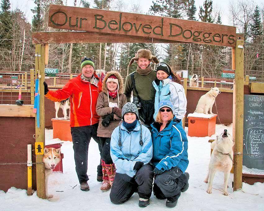 Family posing under 'Out Beloved Doggers' sign with sled dogs relaxing in the background