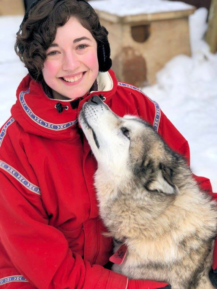 Girl with brown curly hair in a red jacket smiles as she holds a sled dog