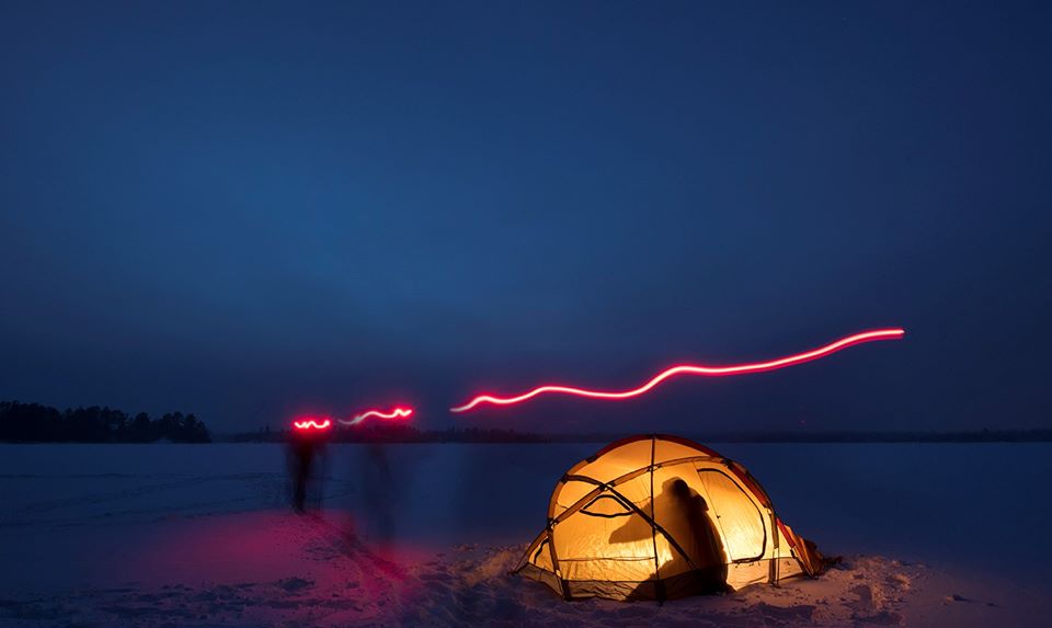 Person in lit tent on frozen lake at night with a long exposed red light making a line