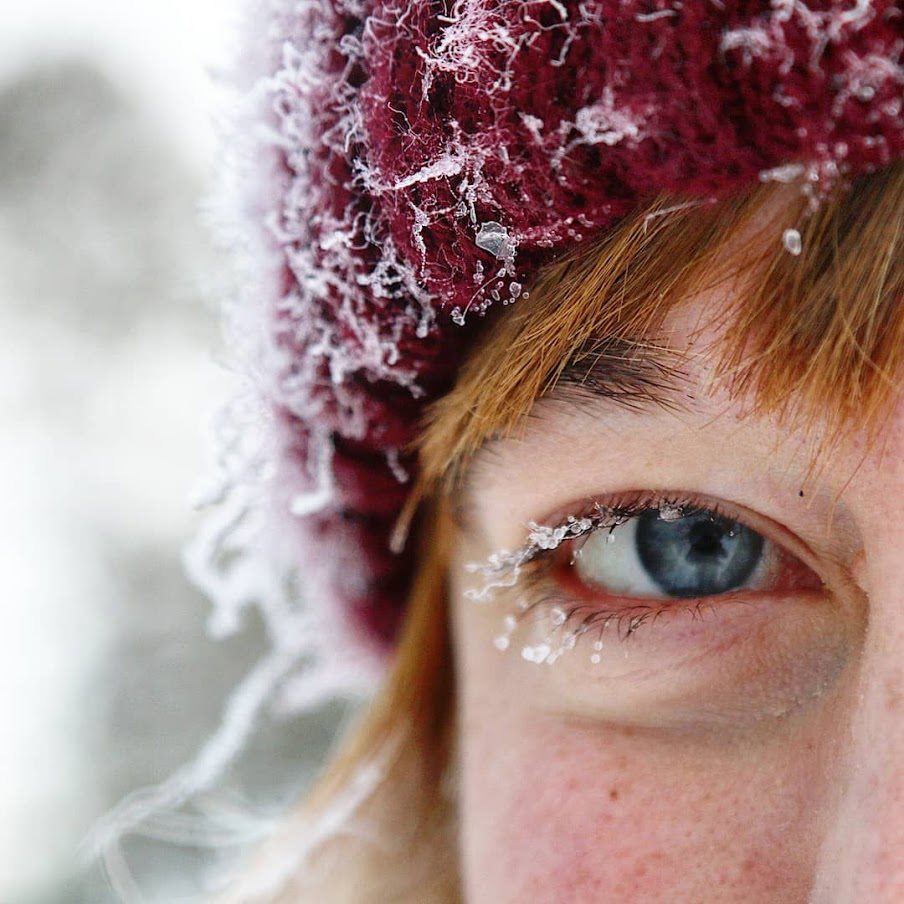Extreme closeup of person with orange hair, red beanie, and blue eyes covered in small snow drops