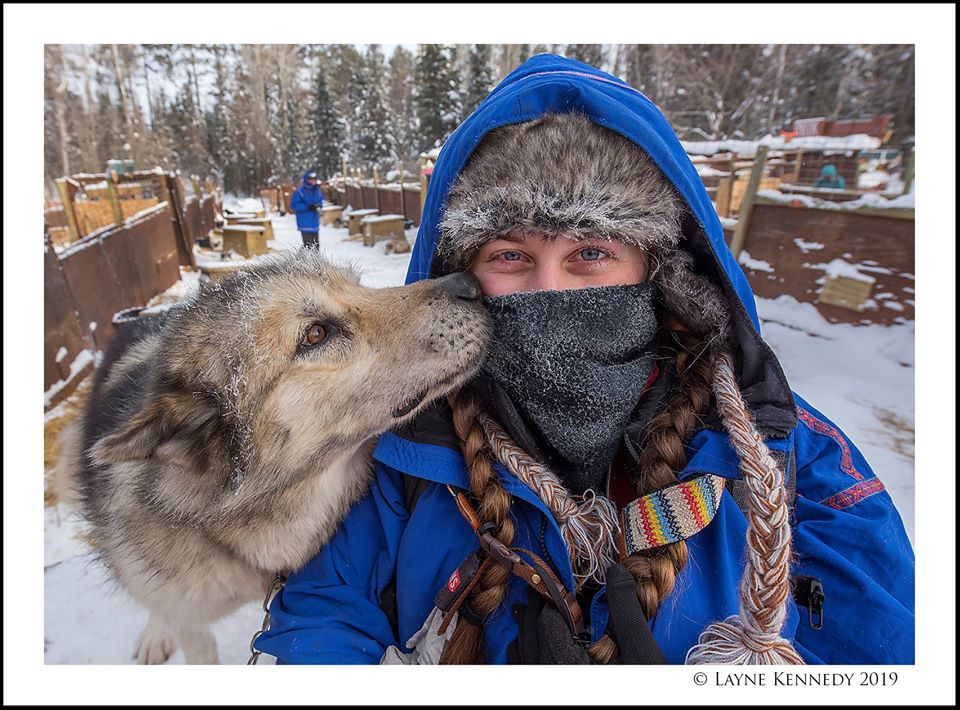Woman in braids and snow gear being kissed by snowdog